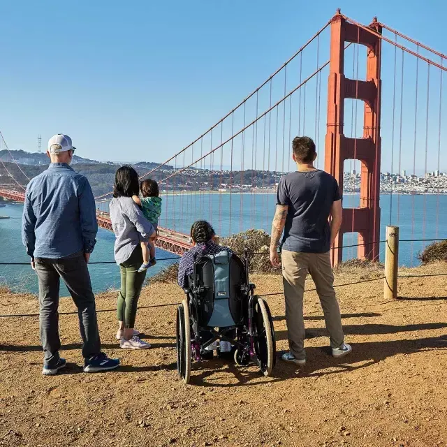 Se ve a un grupo de personas, incluida una persona en silla de ruedas, desde atrás mientras observan el Golden Gate Bridge desde Marin Headlands.